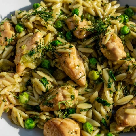 Golden-brown chicken pieces, vibrant green peas, and orzo pasta simmer in a zesty lemon herb sauce inside a warm skillet, ready to serve.