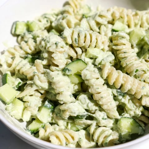 Bright green creamy Green Goddess Pasta Salad in a white bowl, with fresh diced cucumber, spinach, and herbs on a rustic wooden table.