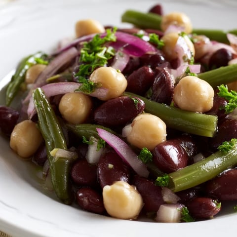 A colorful bowl of Three-Bean Salad with green beans, kidney beans, and chickpeas in a tangy vinaigrette, garnished with fresh parsley and red onion.  