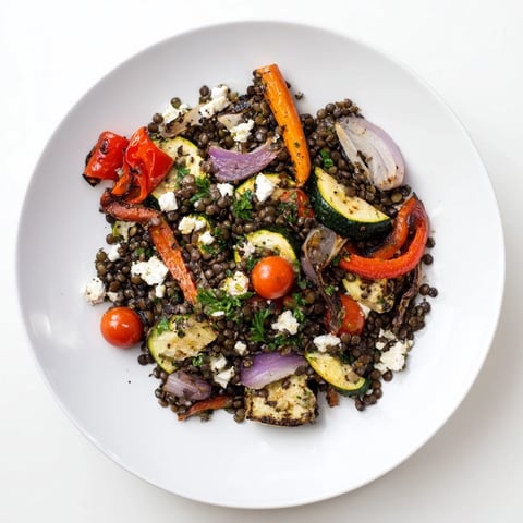 A close-up view of a Black Lentil Salad with Roasted Vegetables, featuring glistening beluga lentils mixed with caramelized red bell peppers and zucchini. Fresh parsley and crumbled feta add color, with a light lemon dressing drizzled over the top.