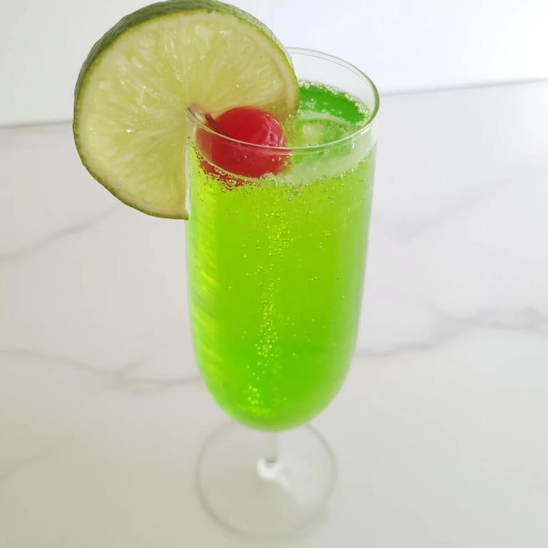 This vibrant Quick Grinch Mimosa Mocktail, a dazzling green non-alcoholic drink, awaits garnishing with cherry slices.